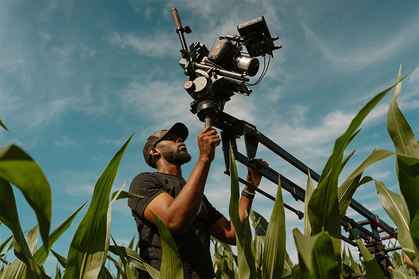 Videographer operating a professional camera rig on a track surrounded by tall plants under a clear blue sky.
