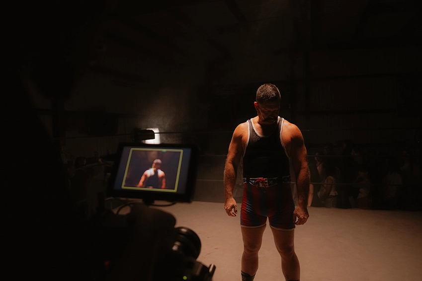 Muscular wrestler standing alone in a dimly lit ring while being filmed, with a camera monitor visible in the foreground and dramatic overhead lighting.