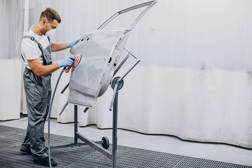 Auto body technician sanding a car door panel in a paint preparation area before refinishing.