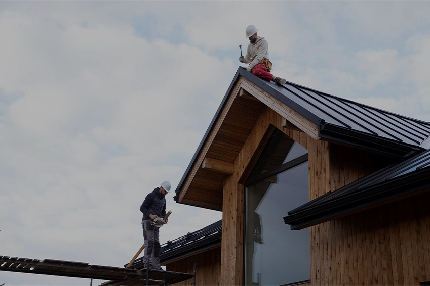 Two roofers installing a black metal roof on a modern wooden house under a cloudy sky.