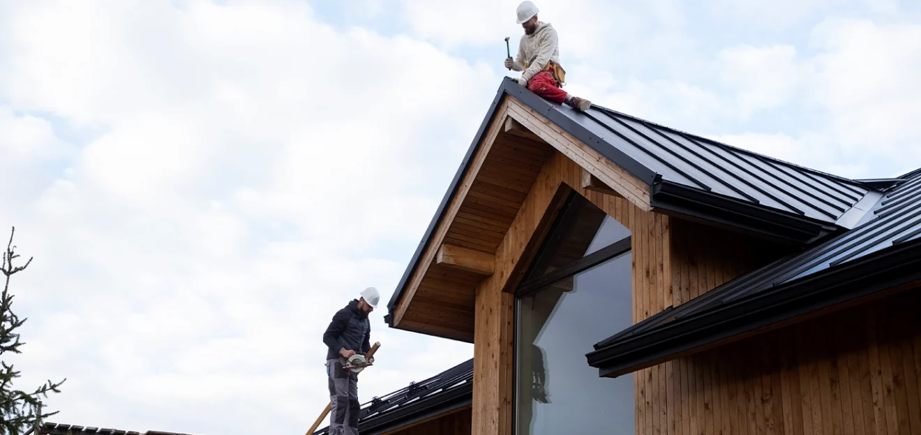Two roofers installing a black metal roof on a modern wooden house under a cloudy sky.