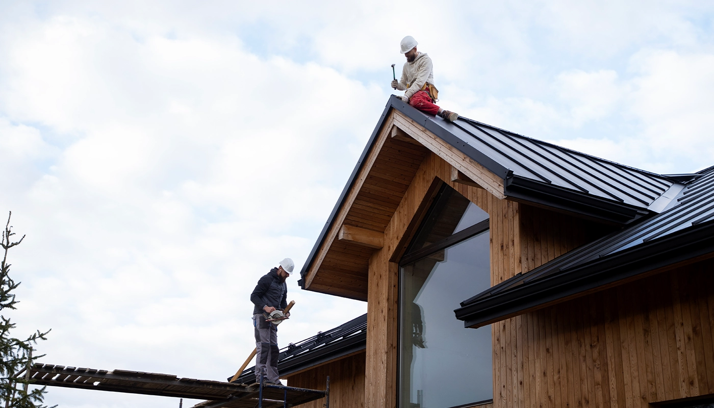 Two roofers installing a black metal roof on a modern wooden house under a cloudy sky.