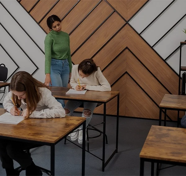 Teacher supervising a classroom of students writing exams at wooden desks in a modern learning environment.
