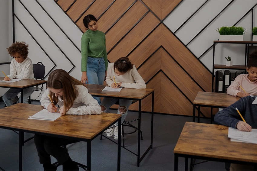Teacher supervising a classroom of students writing exams at wooden desks in a modern learning environment.