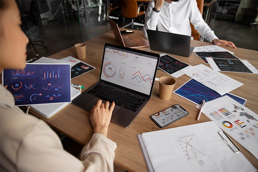 Marketing team analyzing digital performance charts and data analytics on laptops and printed reports in an office meeting.