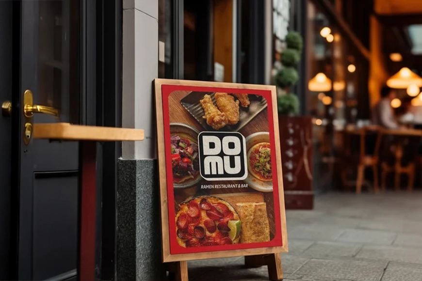 Restaurant sidewalk sign displaying DOMU Ramen branding and food photography — creative brand design and visual marketing.