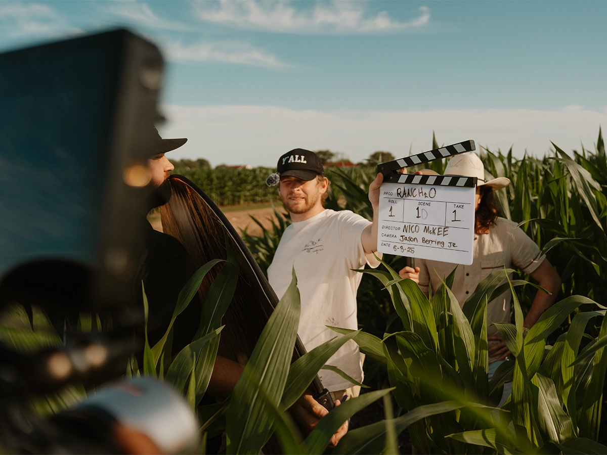Behind-the-scenes photo of a film crew shooting a project titled “RANCH H2O” in a cornfield, featuring a director holding a clapperboard under natural daylight.