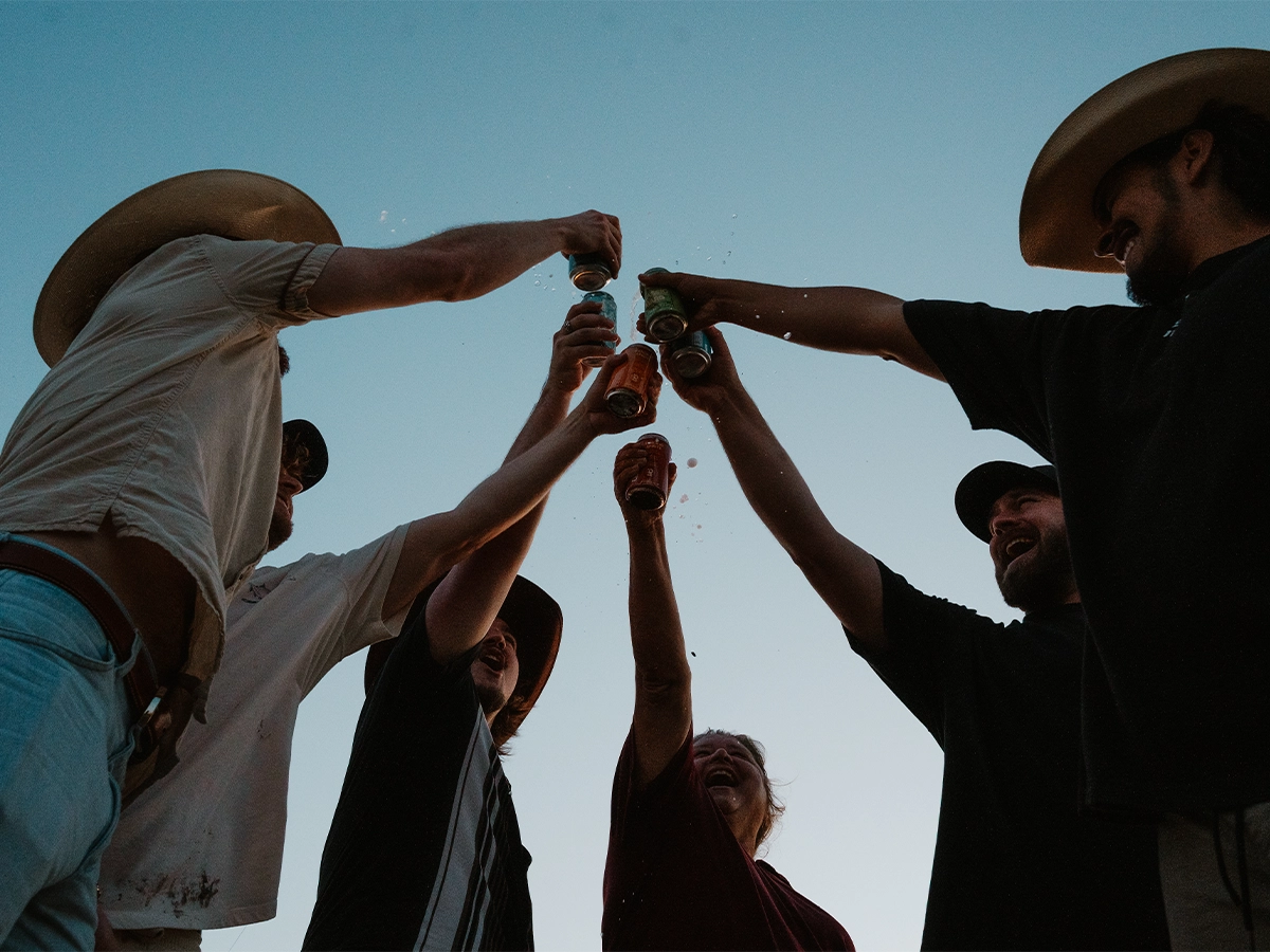 Group of friends raising cans in a celebratory toast under a clear blue sky, symbolizing unity, joy, and shared moments outdoors.