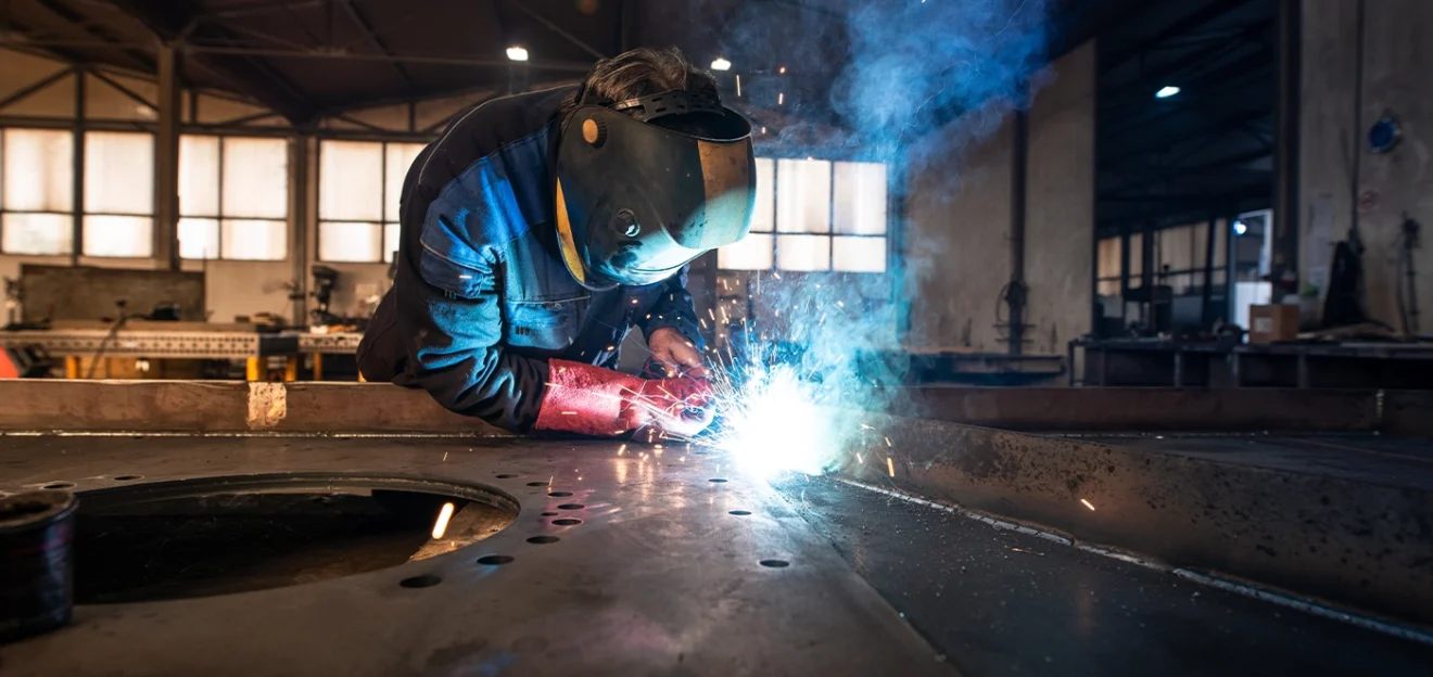 Industrial welder wearing protective gear and gloves working with bright sparks in a metal fabrication workshop.