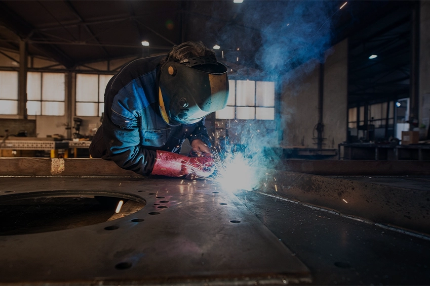 Industrial welder wearing protective gear and gloves working with bright sparks in a metal fabrication workshop.