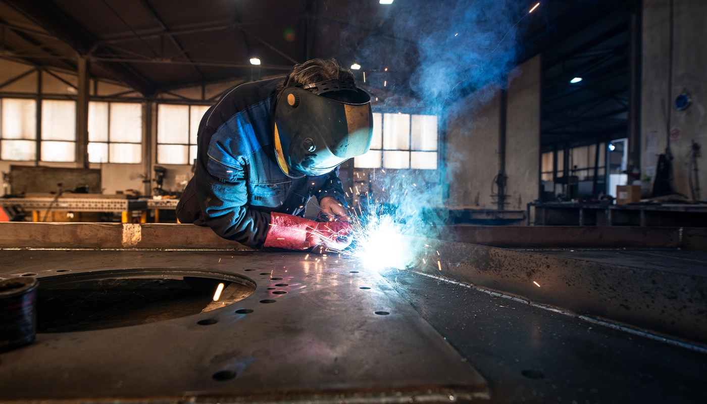 Industrial welder wearing protective gear and gloves working with bright sparks in a metal fabrication workshop.