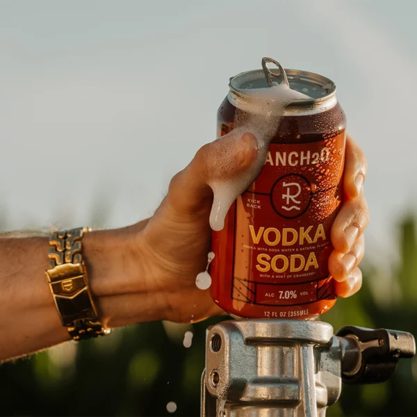 Hand holding an open can of Ranch2O Vodka Soda with foam spilling over the top during a warm outdoor setting.