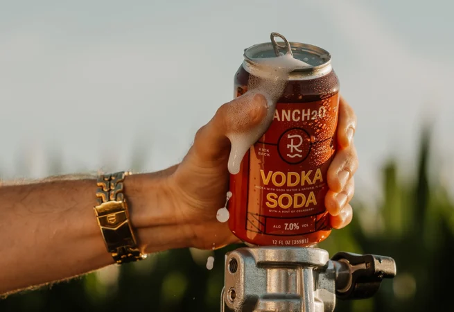 Hand holding an open can of Ranch2O Vodka Soda with foam spilling over the top during a warm outdoor setting.