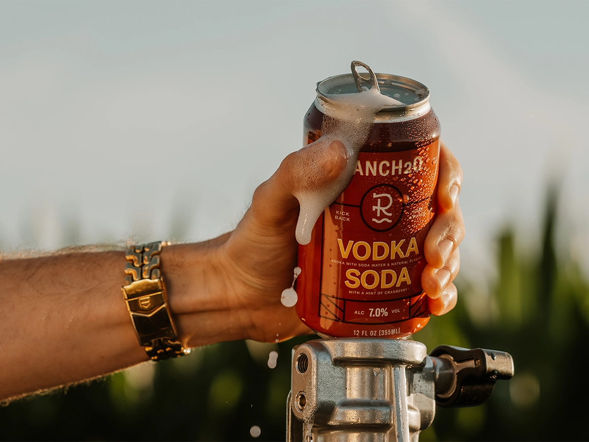 Hand holding an open can of Ranch2O Vodka Soda with foam spilling over the top during a warm outdoor setting.