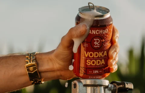 Hand holding an open can of Ranch2O Vodka Soda with foam spilling over the top during a warm outdoor setting.
