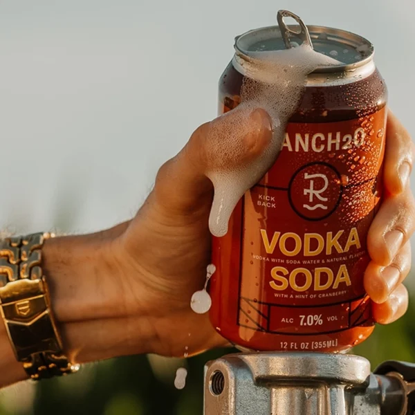 Hand holding an open can of Ranch2O Vodka Soda with foam spilling over the top during a warm outdoor setting.