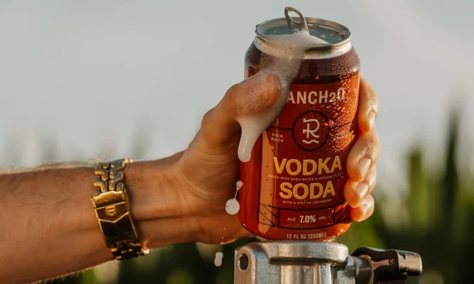 Hand holding an open can of Ranch2O Vodka Soda with foam spilling over the top during a warm outdoor setting.