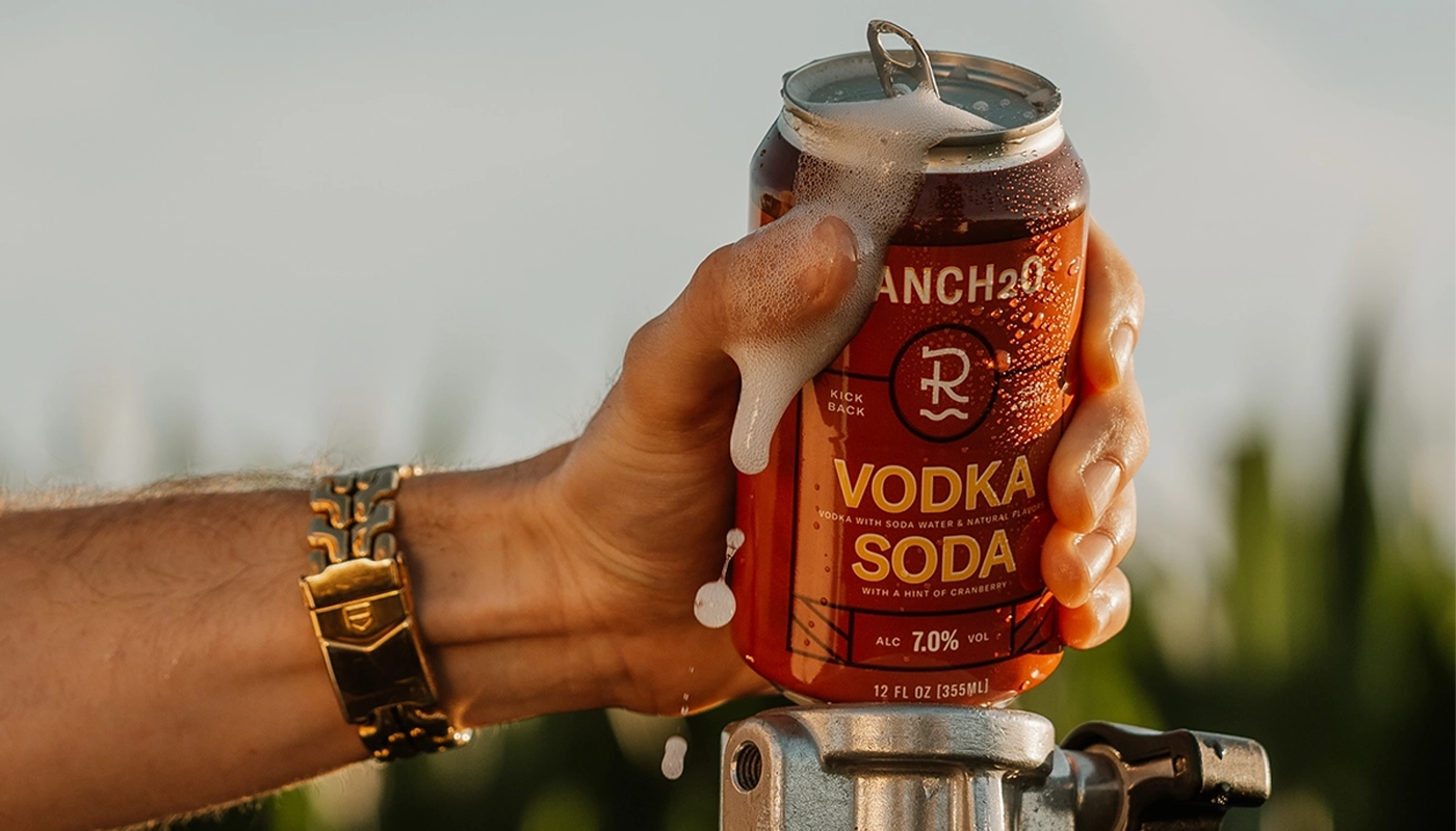 Hand holding an open can of Ranch2O Vodka Soda with foam spilling over the top during a warm outdoor setting.