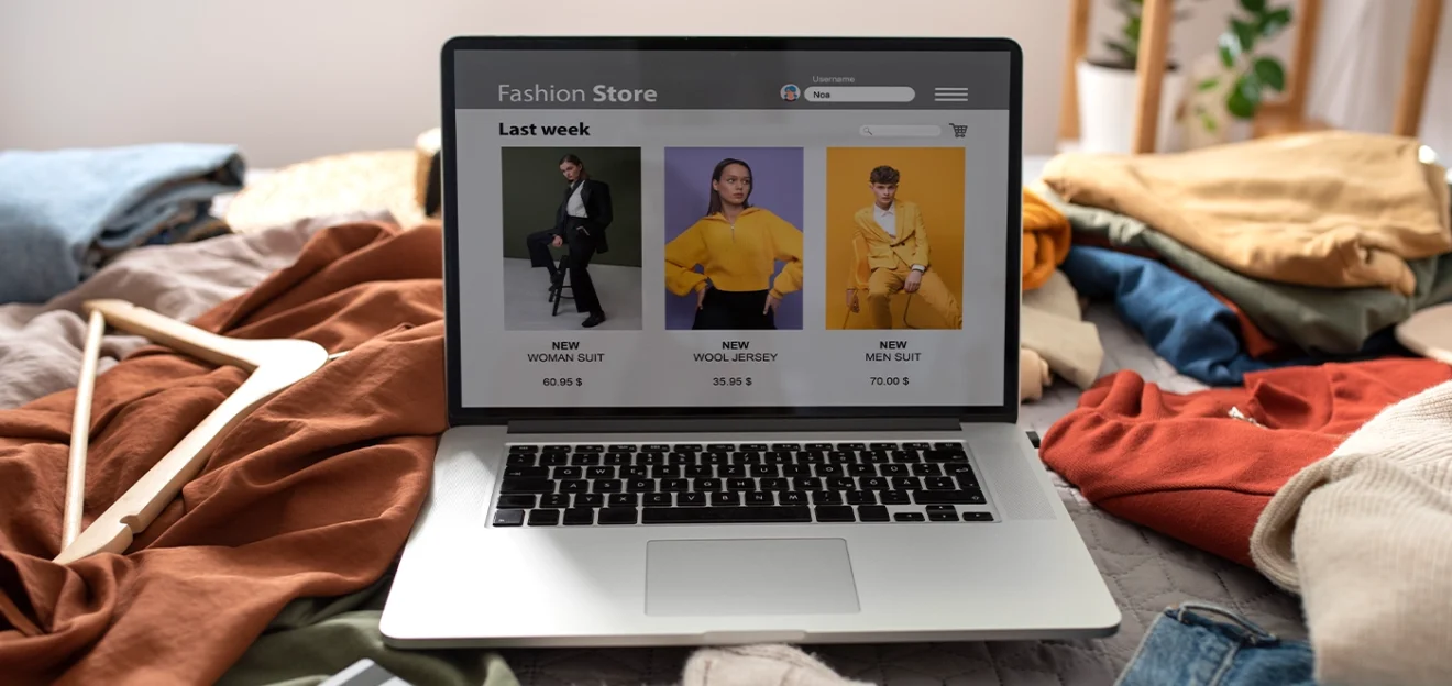 Laptop showing an online fashion store with new clothing collections, surrounded by casual clothes and accessories on a bed.