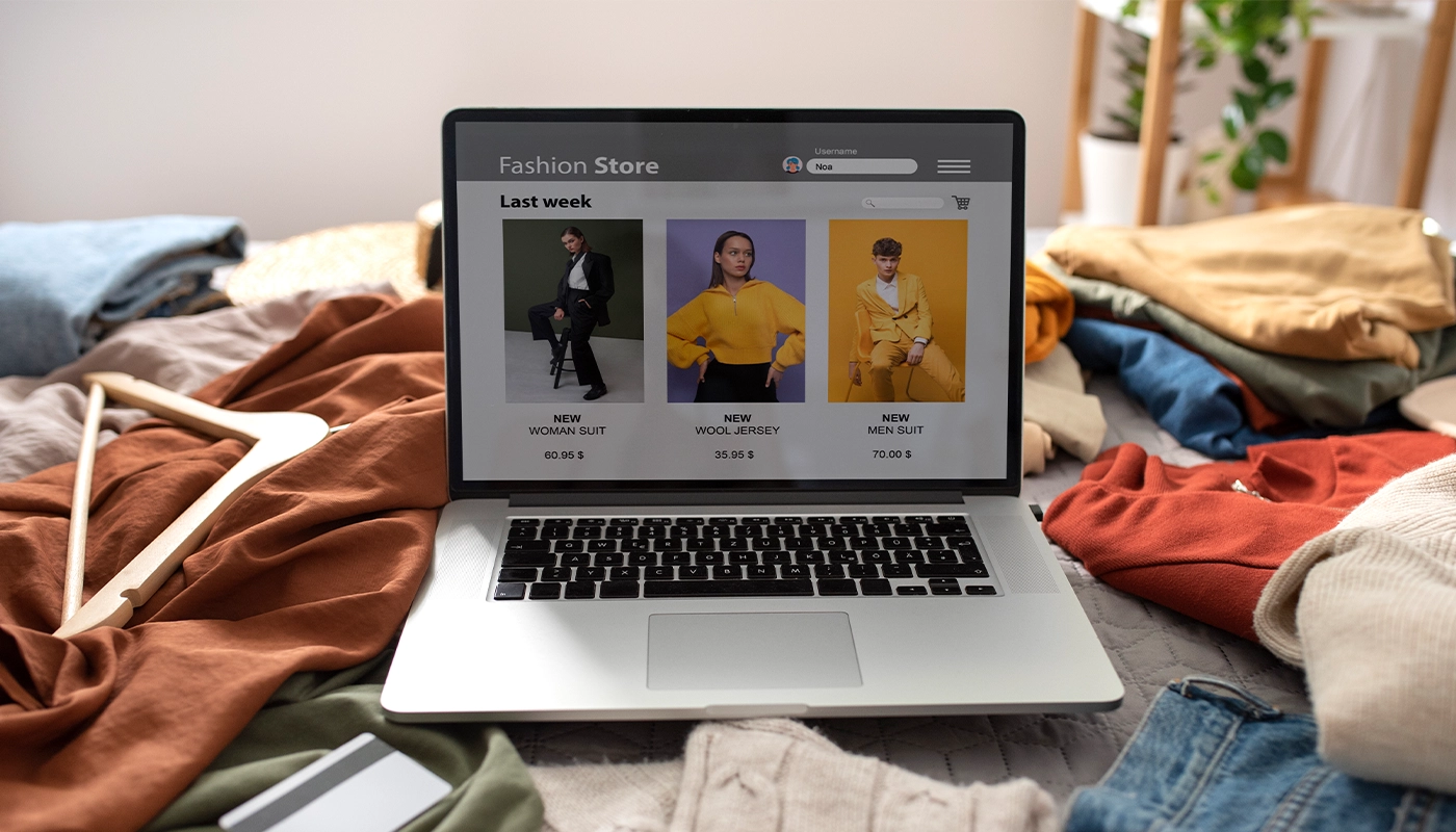 Laptop showing an online fashion store with new clothing collections, surrounded by casual clothes and accessories on a bed.