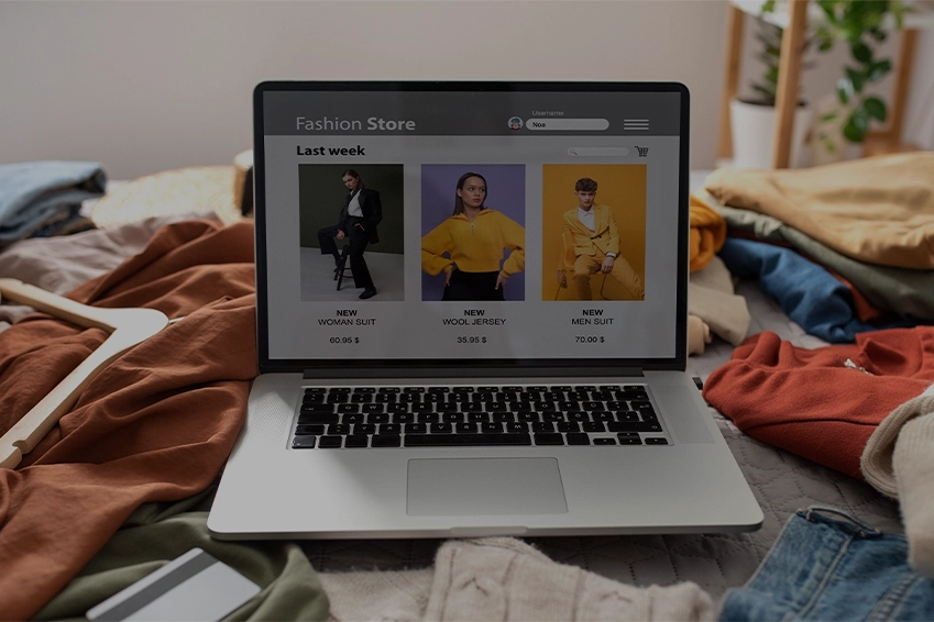 Laptop showing an online fashion store with new clothing collections, surrounded by casual clothes and accessories on a bed.