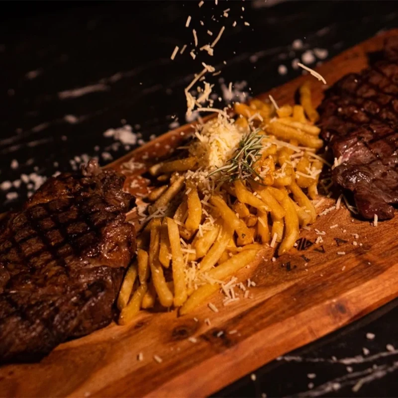 Gourmet steak dinner served on a wooden board with crispy fries topped with shredded cheese and rosemary, presented under warm lighting for a fine-dining experience.