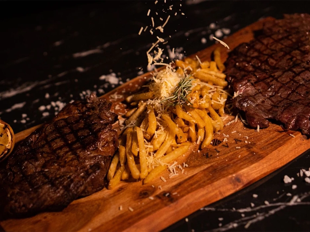 Gourmet steak dinner served on a wooden board with crispy fries topped with shredded cheese and rosemary, presented under warm lighting for a fine-dining experience.