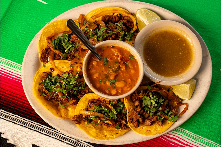 Plate of street-style tacos topped with seasoned meat, onions, and cilantro, served with beans, green salsa, and lime wedges on a colorful Mexican tablecloth.