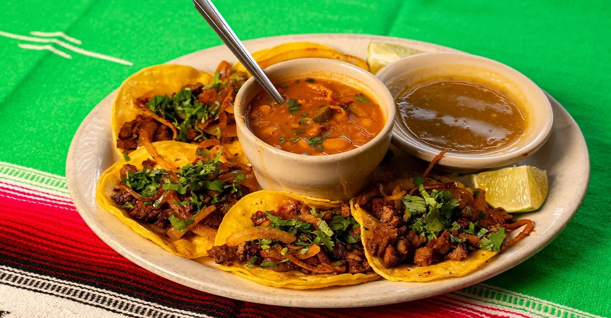Wide shot of a plate of authentic street tacos with seasoned meat, onions, and cilantro, served with beans, green salsa, and lime wedges on a colorful Mexican tablecloth.