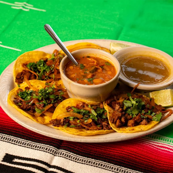 Wide shot of a plate of authentic street tacos with seasoned meat, onions, and cilantro, served with beans, green salsa, and lime wedges on a colorful Mexican tablecloth.