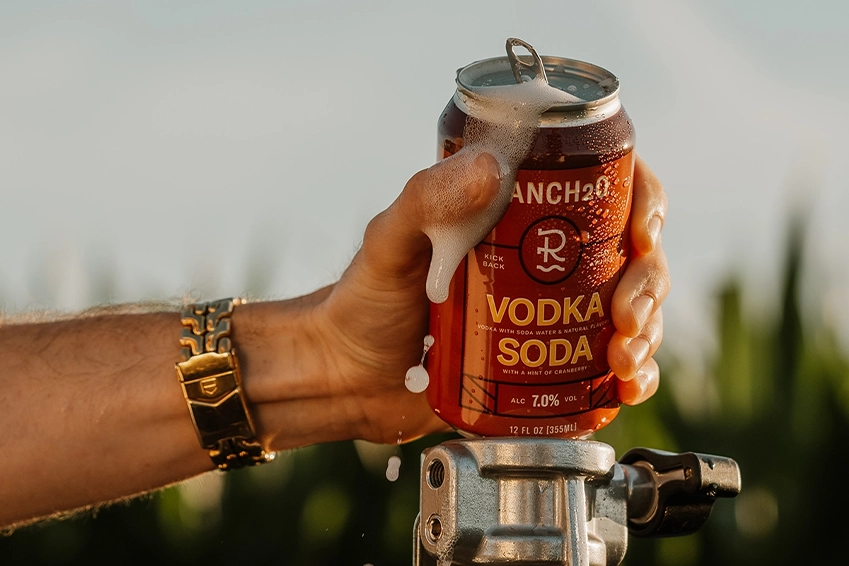 Hand holding an open can of Ranch2O Vodka Soda with foam spilling over the top during a warm outdoor setting.