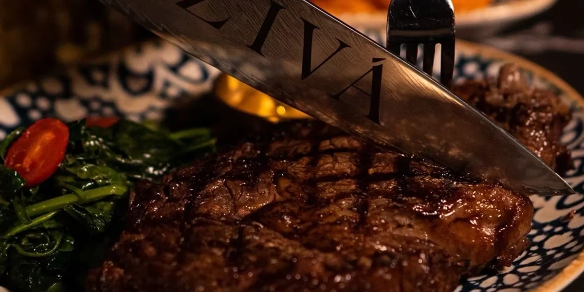 Close-up of a juicy grilled steak being sliced with a knife engraved with the word “ZIVÁ,” served with sautéed greens and cherry tomatoes on a patterned plate.
