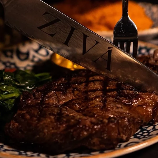 Close-up of a juicy grilled steak being sliced with a knife engraved with the word “ZIVÁ,” served with sautéed greens and cherry tomatoes on a patterned plate.