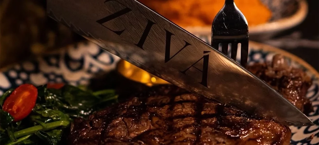 Close-up of a juicy grilled steak being sliced with a knife engraved with the word “ZIVÁ,” served with sautéed greens and cherry tomatoes on a patterned plate.