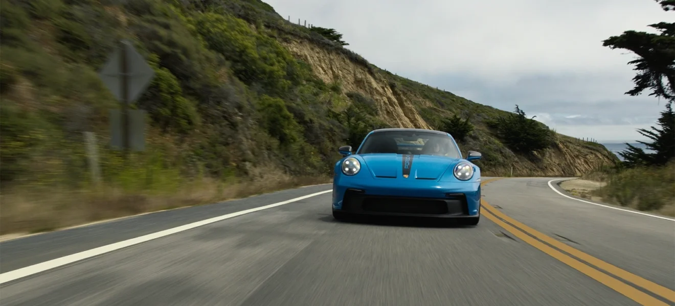 Baby blue Porsche 911 GT3 driving along a winding coastal road with cliffs and greenery in the background.