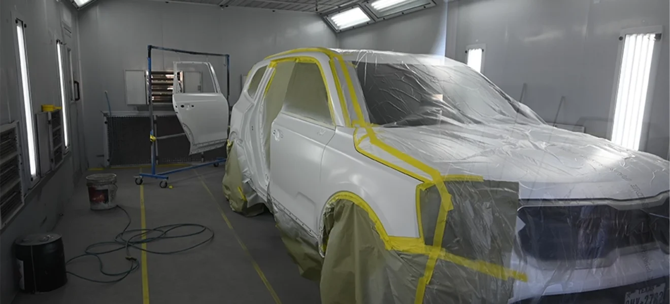 A white SUV is masked with plastic and tape inside a professional automotive paint booth, with bright overhead lights and detached car panels prepared for refinishing at an auto body shop.