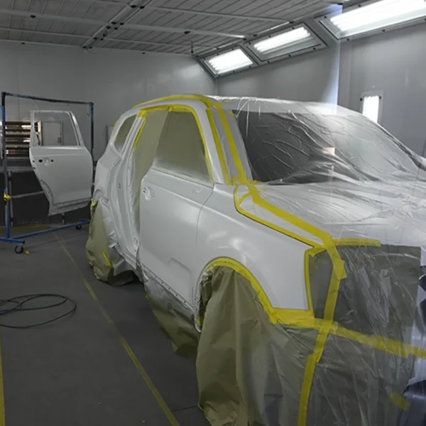 A white SUV is masked with plastic and tape inside a professional automotive paint booth, with bright overhead lights and detached car panels prepared for refinishing at an auto body shop.