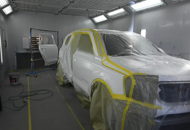 A white SUV is masked with plastic and tape inside a professional automotive paint booth, with bright overhead lights and detached car panels prepared for refinishing at an auto body shop.
