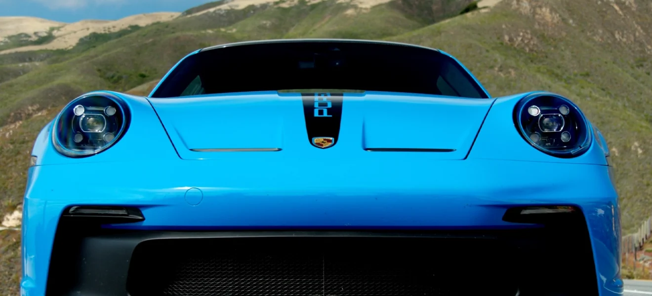 Baby blue Porsche 911 GT3 driving along a winding coastal road with cliffs and greenery in the background.