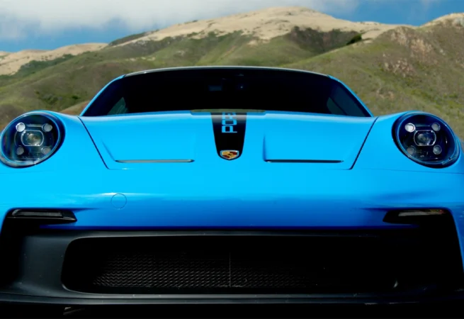 Baby blue Porsche 911 GT3 driving along a winding coastal road with cliffs and greenery in the background.